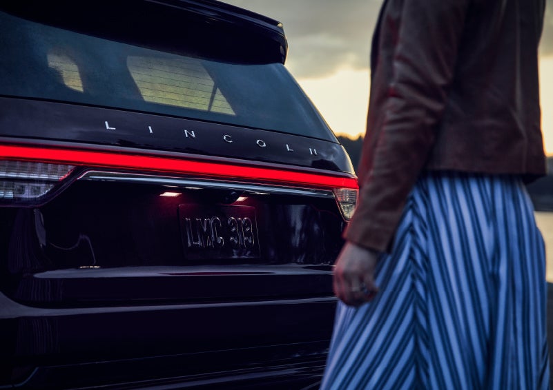 A person is shown near the rear of a 2024 Lincoln Aviator® SUV as the Lincoln Embrace illuminates the rear lights | Dutch Miller Lincoln of Ashland in Ashland KY