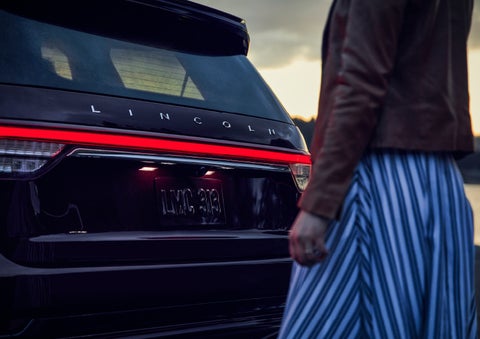 A person is shown near the rear of a 2024 Lincoln Aviator® SUV as the Lincoln Embrace illuminates the rear lights | Dutch Miller Lincoln of Ashland in Ashland KY